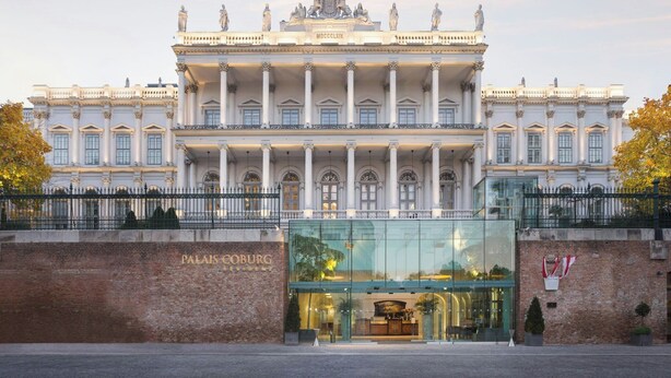 Un edificio imponente de arquitectura clásica, que presenta una entrada central con columnas, un balcón amplio y una placa con la inscripción 'Palais Coburg'.