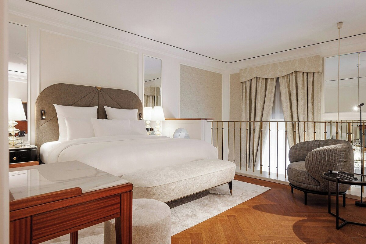 A hotel room with a large bed, a desk, a chair, and a balcony with curtains.