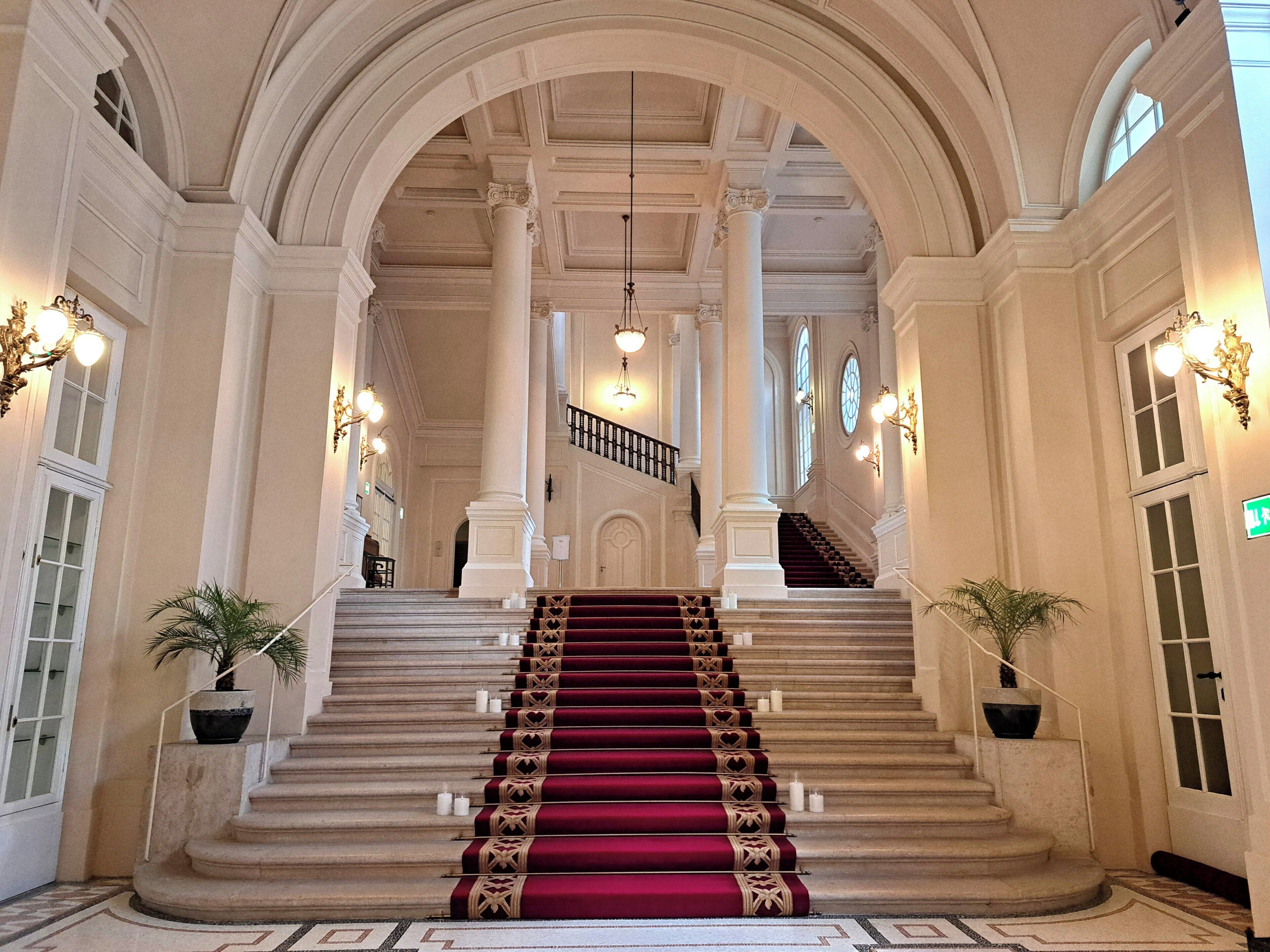A grand staircase with a red carpet, flanked by columns and potted plants, leading to an upper level.