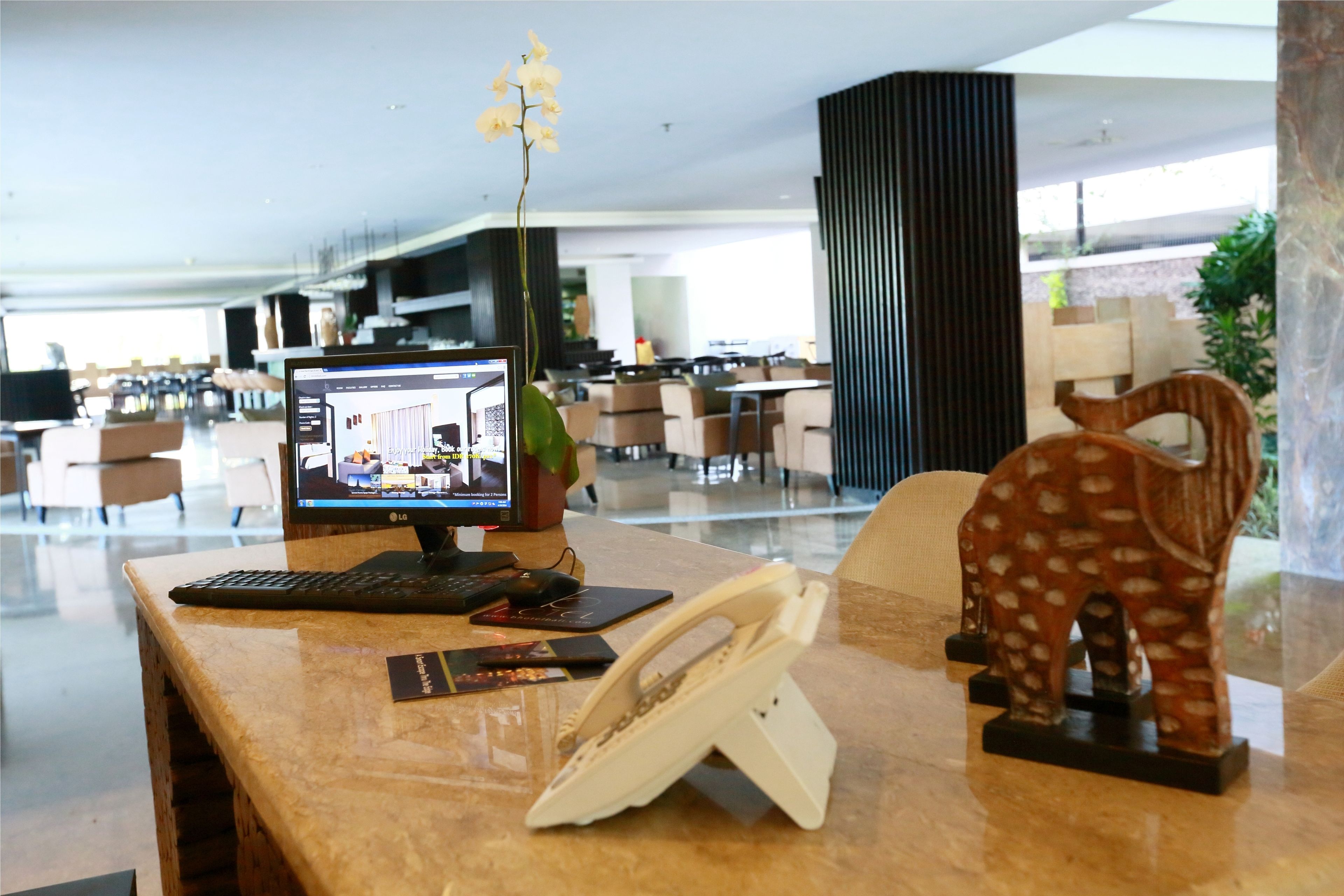 A reception desk with a computer monitor, telephone, and decorative items in a modern lobby with seating and a plant.
