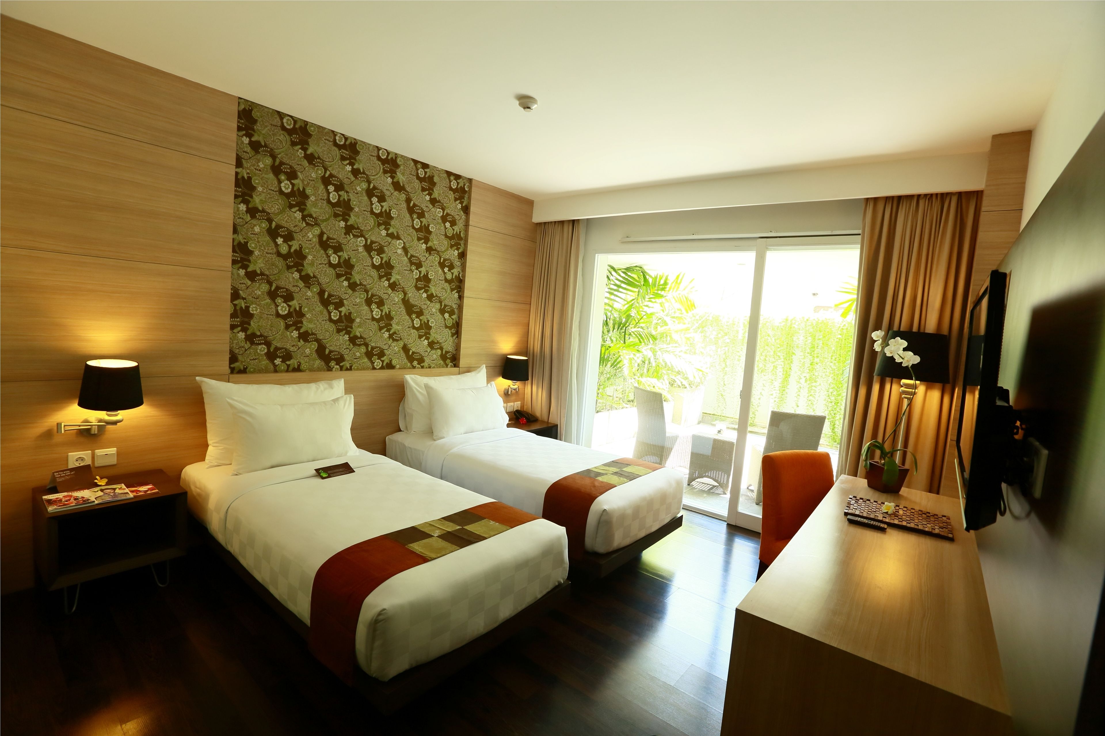 A hotel room with two beds, a desk, a TV, and a view of greenery outside.