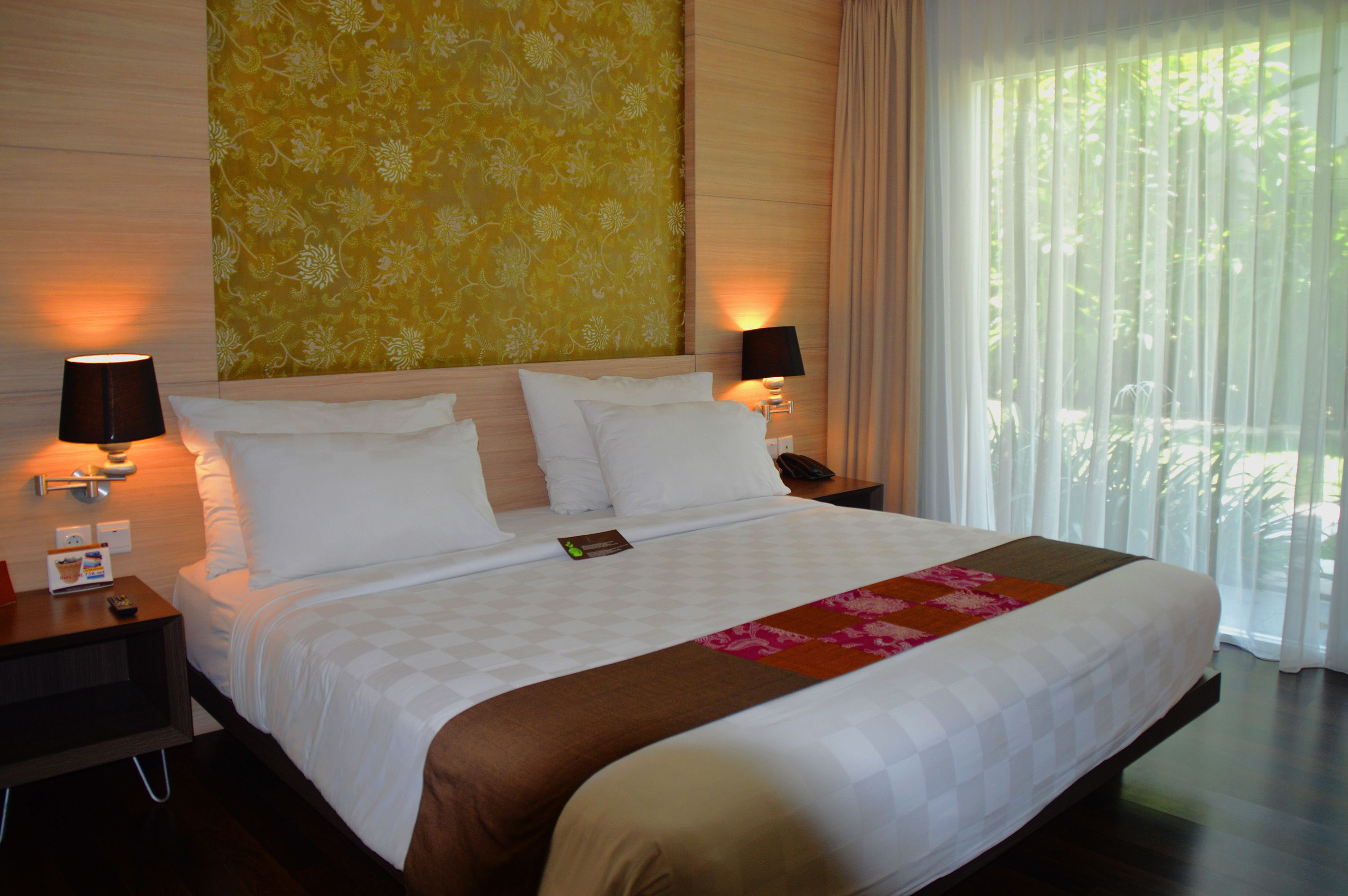 A hotel room with a large bed, two bedside tables, a nightstand, a lamp, and a view of greenery through sheer curtains.