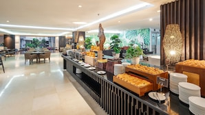 Uma área de buffet com diversos alimentos, um espaço de estar com mesas e cadeiras, e um mural com folhagens tropicais ao fundo.