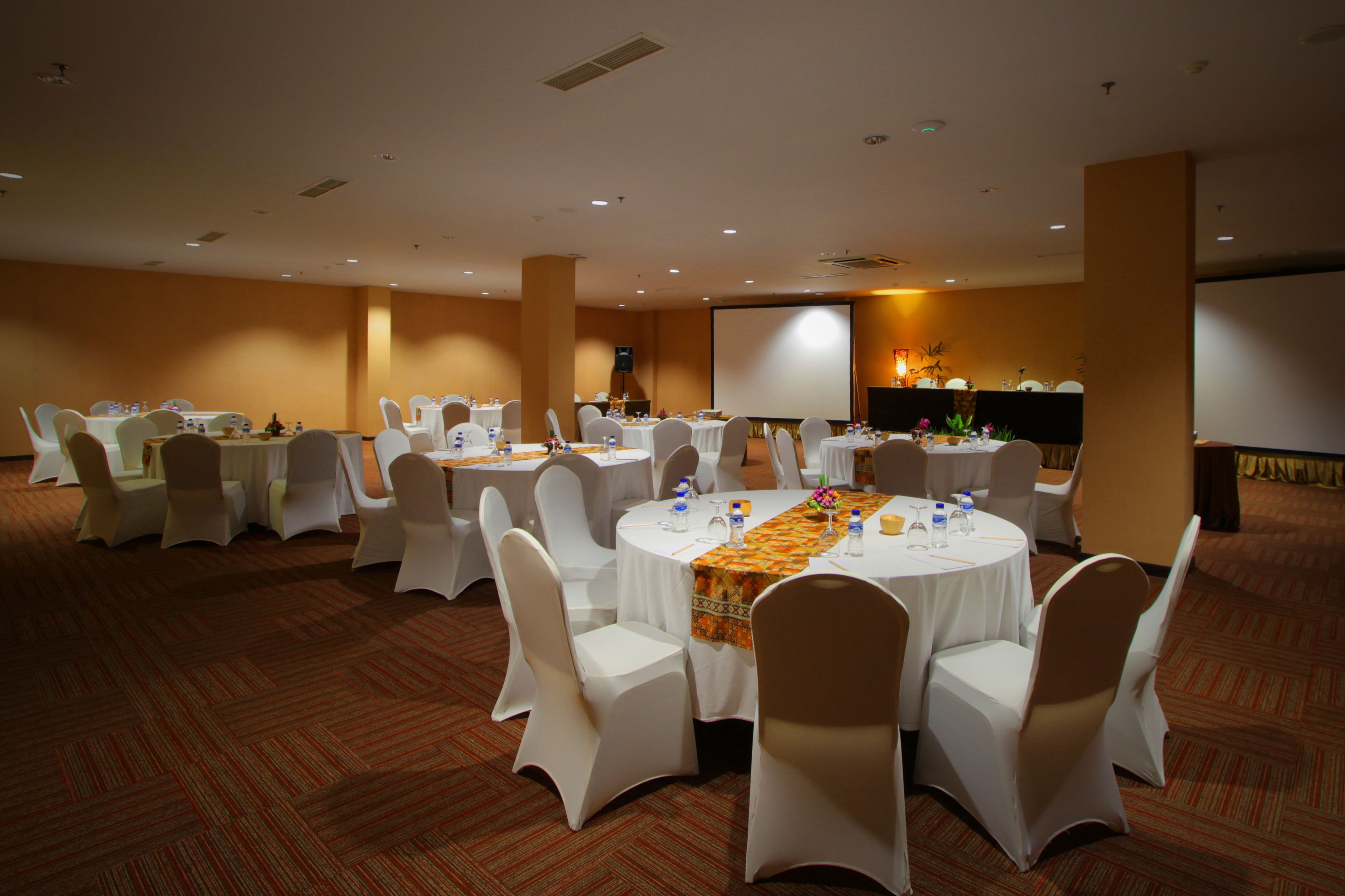 Un salón de conferencias con varias mesas redondas, sillas blancas y una pantalla de proyector.