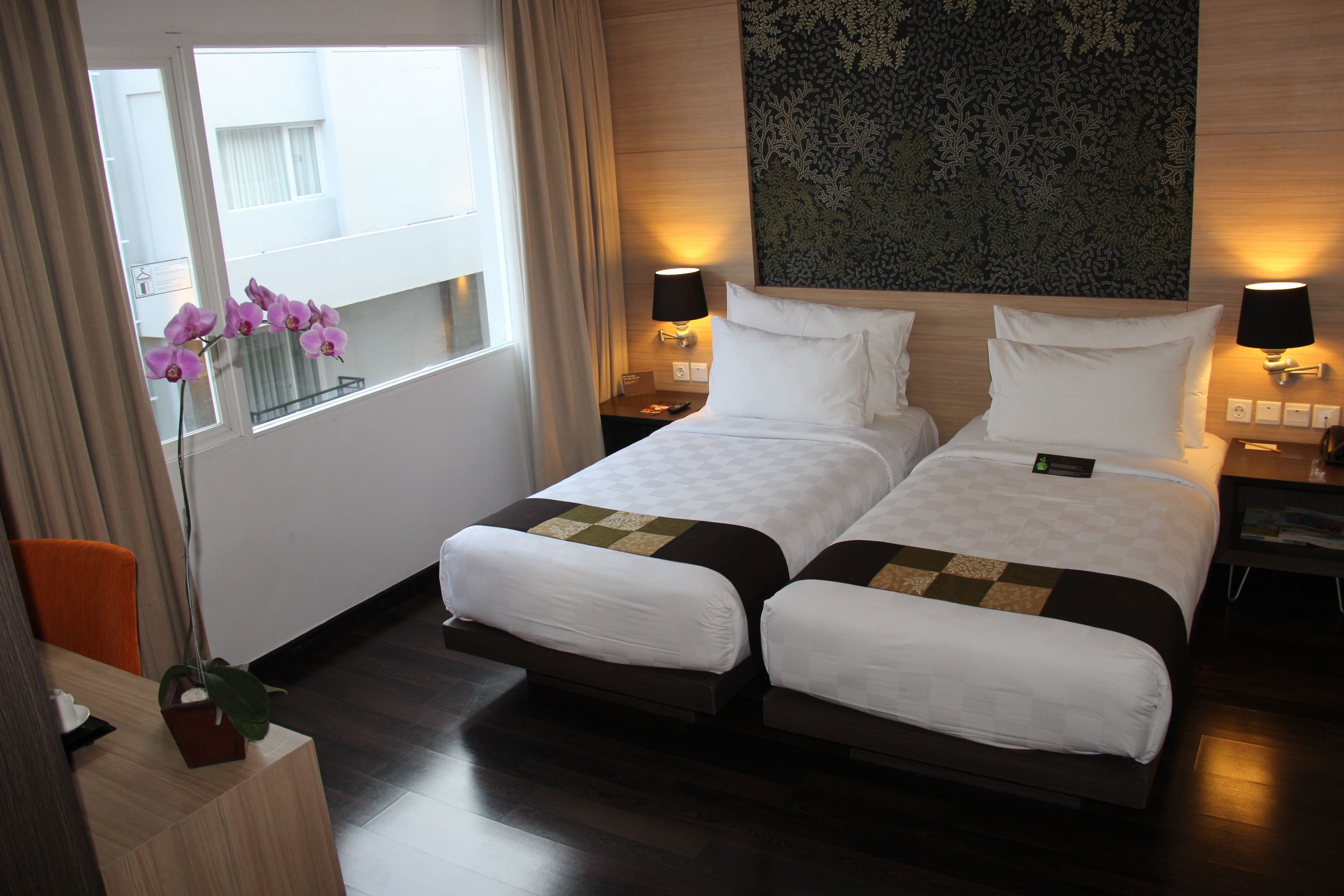 A hotel room with two beds, a large window with curtains, a potted plant, and a decorative wall piece.