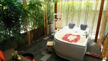 A spa area with a large bathtub filled with rose petals, surrounded by lush greenery and a small seating area with a flower arrangement.