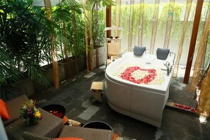 A spa area with a large bathtub filled with rose petals, surrounded by lush greenery and a small seating area with a flower arrangement.