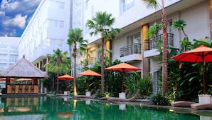 Uma área de piscina de hotel com espreguiçadeiras e guarda-sóis, cercada por palmeiras e um espaço de bar com telhado de palha.