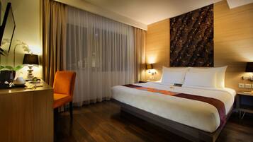 A hotel room with a large bed, a desk with a chair, a desk lamp, a potted plant, and a wall-mounted TV.