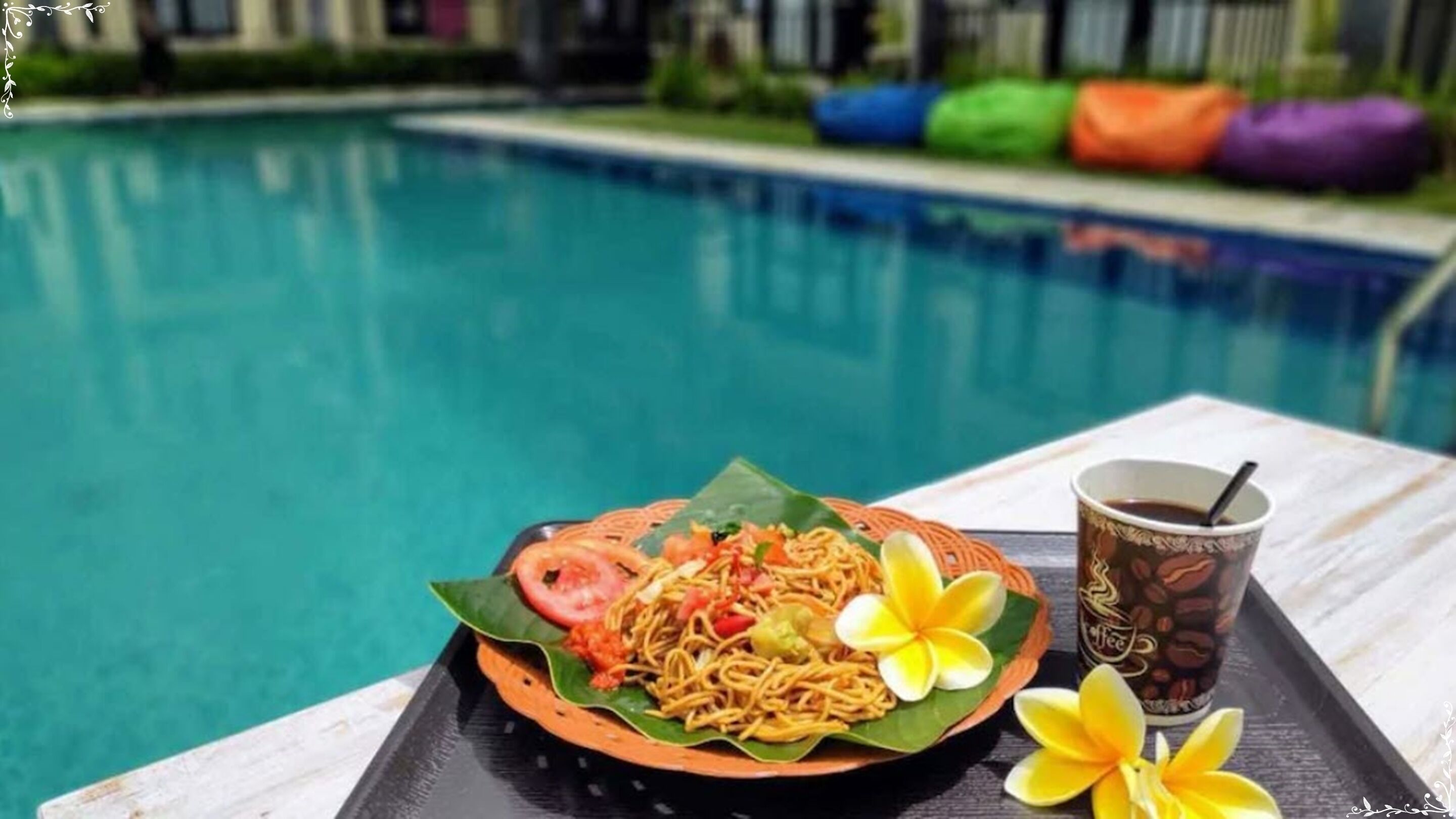 Une table près d’une piscine, avec une assiette de nouilles, une tasse de café et une fleur.