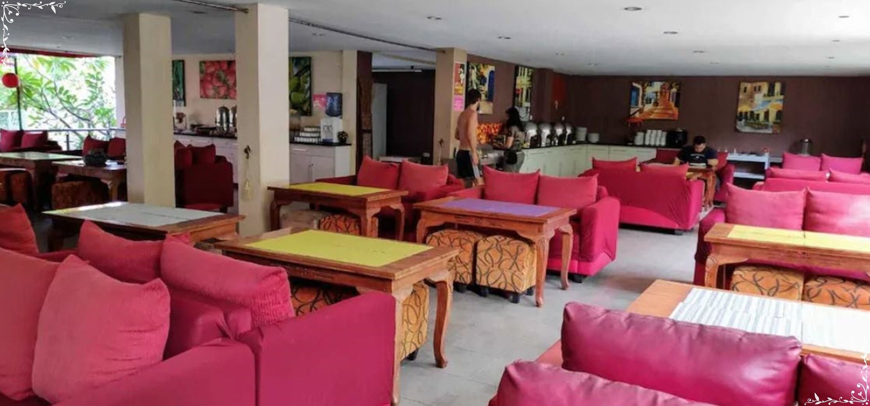 Un restaurante con sofás tapizados en rojo y mesas de madera, una zona de cocina y obras de arte en las paredes.