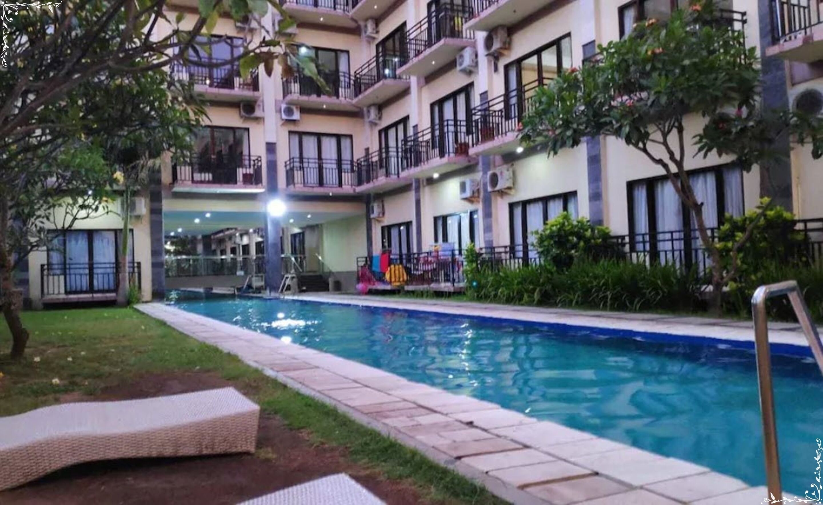 A swimming pool with lounge chairs, surrounded by a multi-story building with balconies and air conditioning units.