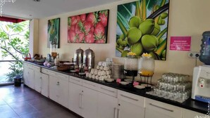 Un comptoir pour le petit-déjeuner avec des machines à café, des théières et un distributeur d’eau. Des tableaux représentant des fruits sont accrochés au mur, ainsi qu’un panneau avec du texte.