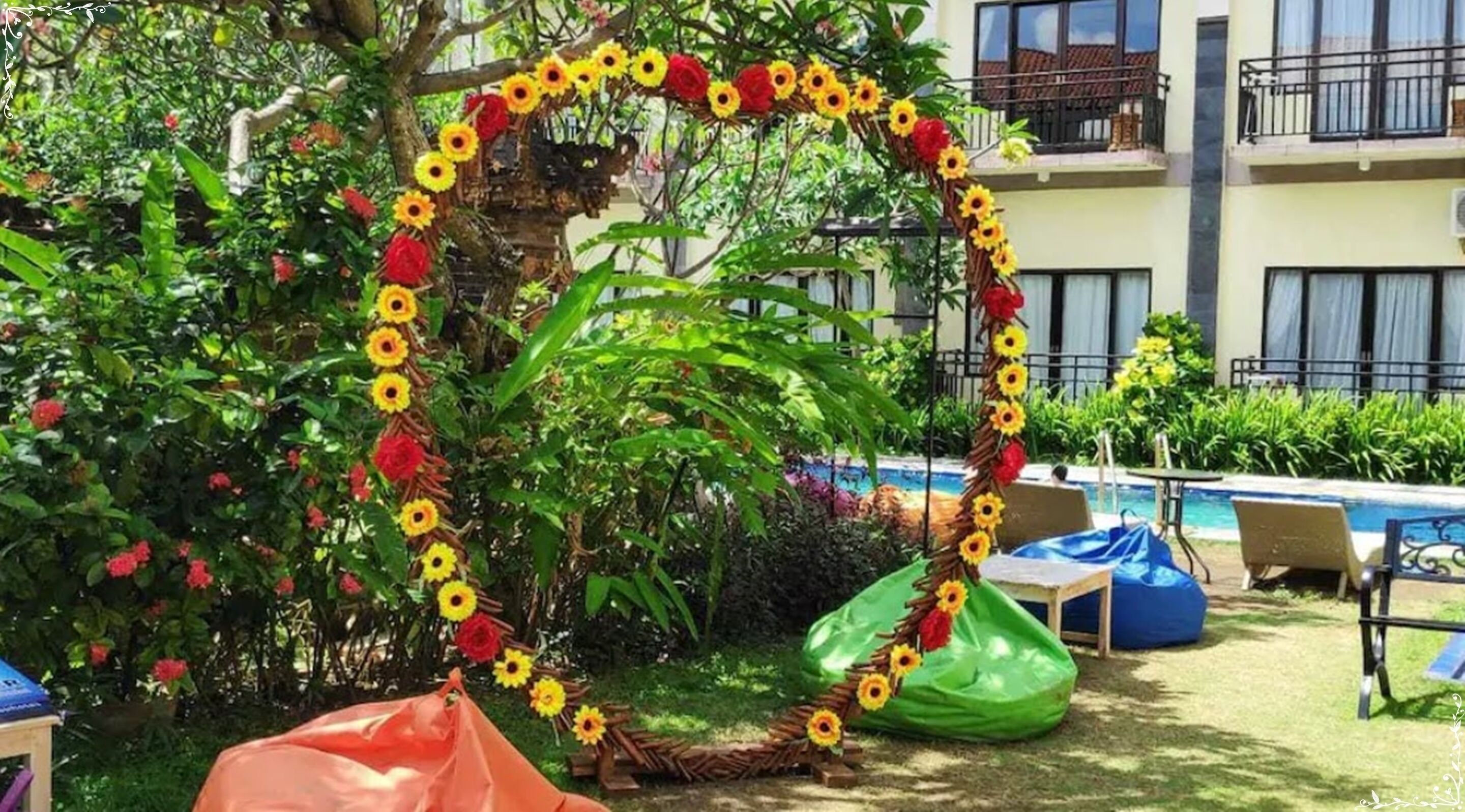 A poolside area with a decorative heart-shaped arrangement of sunflowers and red flowers, surrounded by lush greenery and a building in the background.