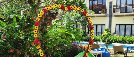A poolside area with a decorative heart-shaped arrangement of sunflowers and red flowers, surrounded by lush greenery and a building in the background.