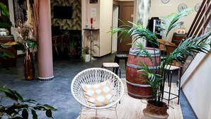 A spacious lobby with a high ceiling, hanging wicker lamps, a variety of seating options including a white wicker chair, a round table, and a woven bench. There are potted plants and a large wooden barrel in the center. - Auberge de jeunesse HI Bordeaux-Blanquefort (Ex Otellia) (Blanquefort)