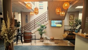 A modern interior with a staircase, wicker furniture, a large wooden barrel, and a mounted TV displaying a scenic view. - Auberge de jeunesse HI Bordeaux-Blanquefort (Ex Otellia) (Blanquefort)