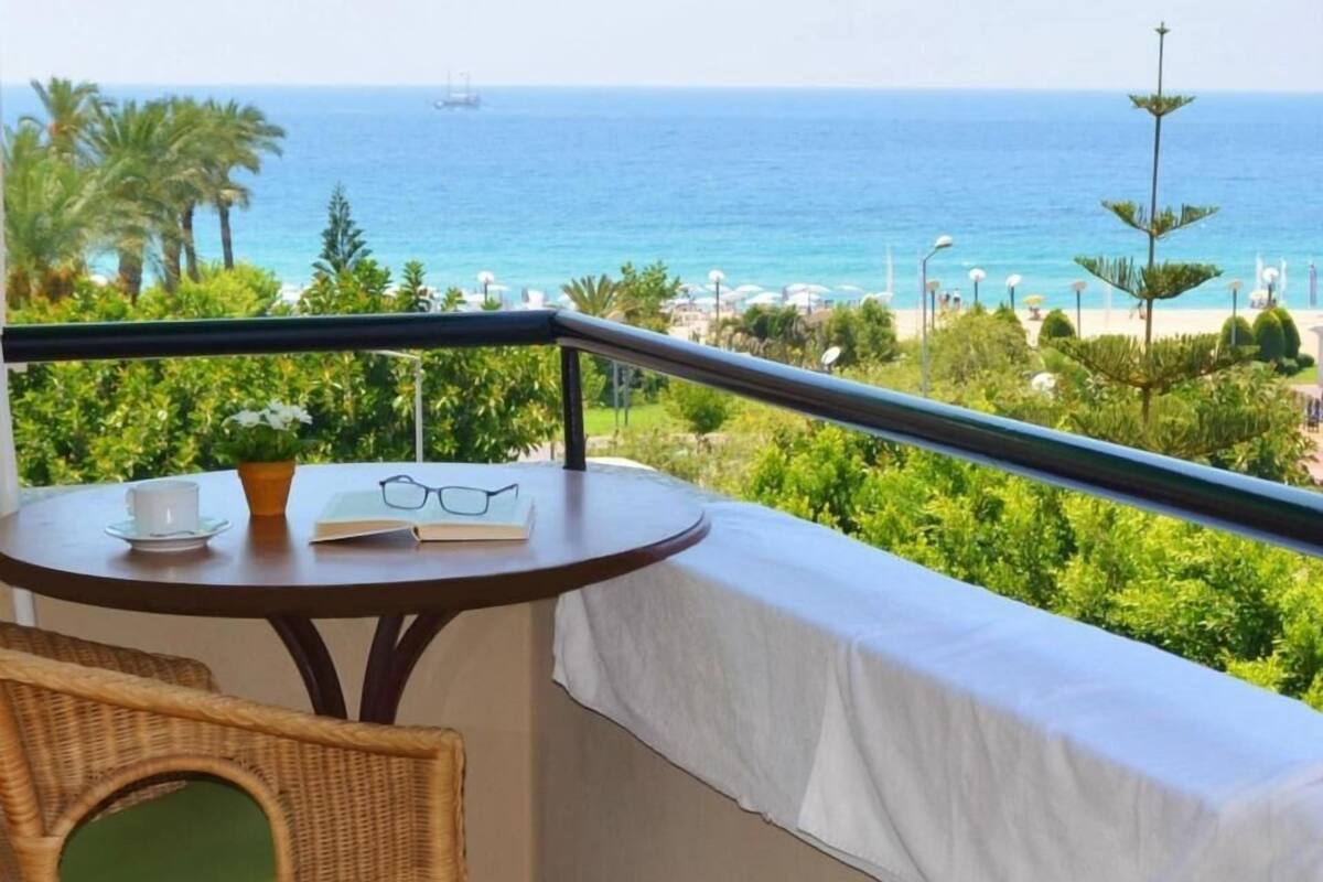 Een balkon met uitzicht op zee, een tafel met een boek en glazen, en een rieten stoel.