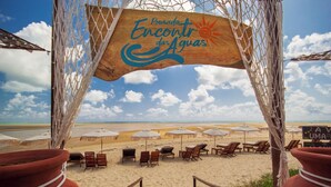 Una playa con un cartel de madera que dice "Pousada Encantada das Aguias". Hay varios sombrillas y tumbonas dispuestas sobre la arena.