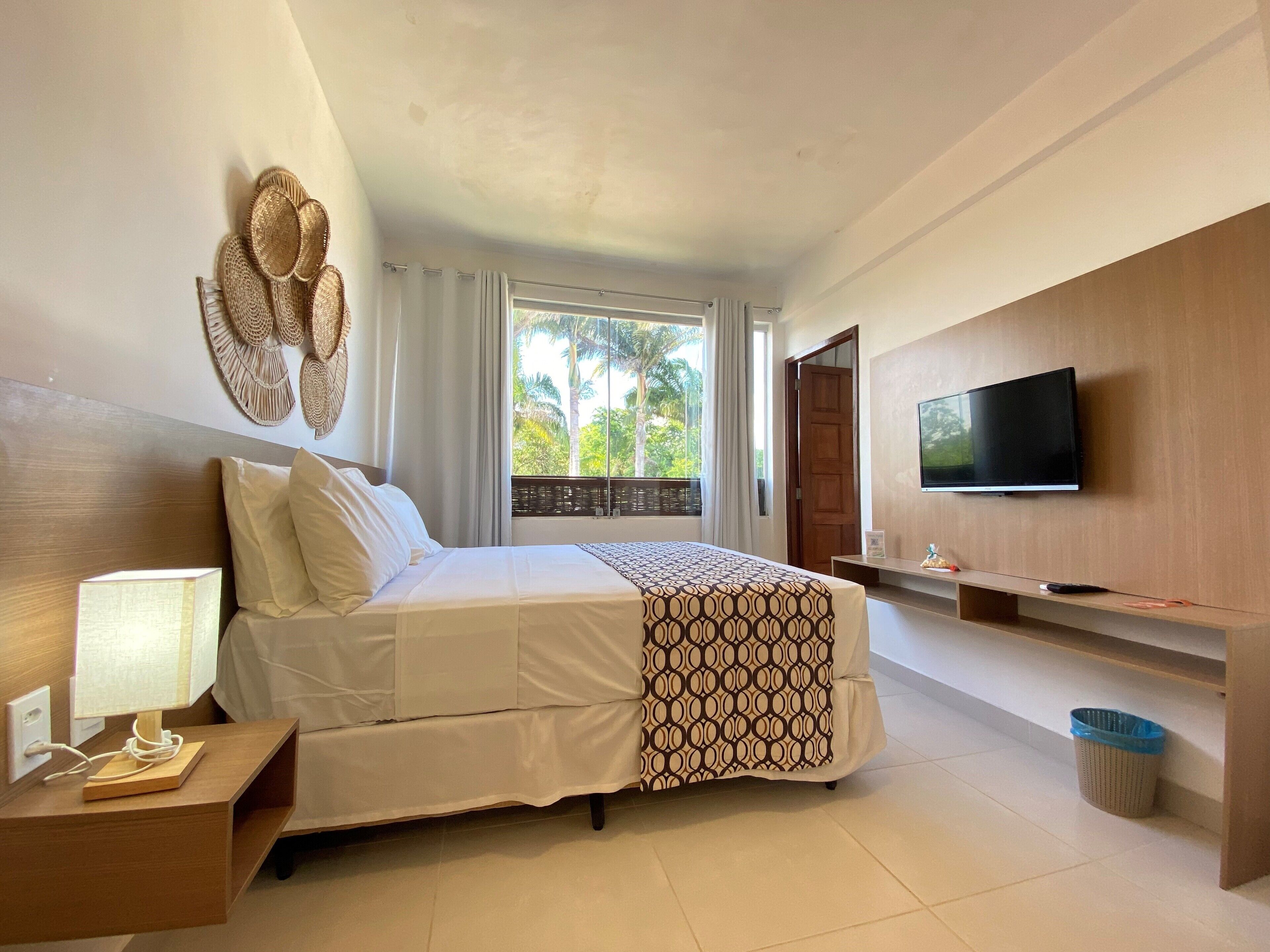 A hotel room with a bed, a TV, a bedside table with a lamp, and a view of greenery outside.