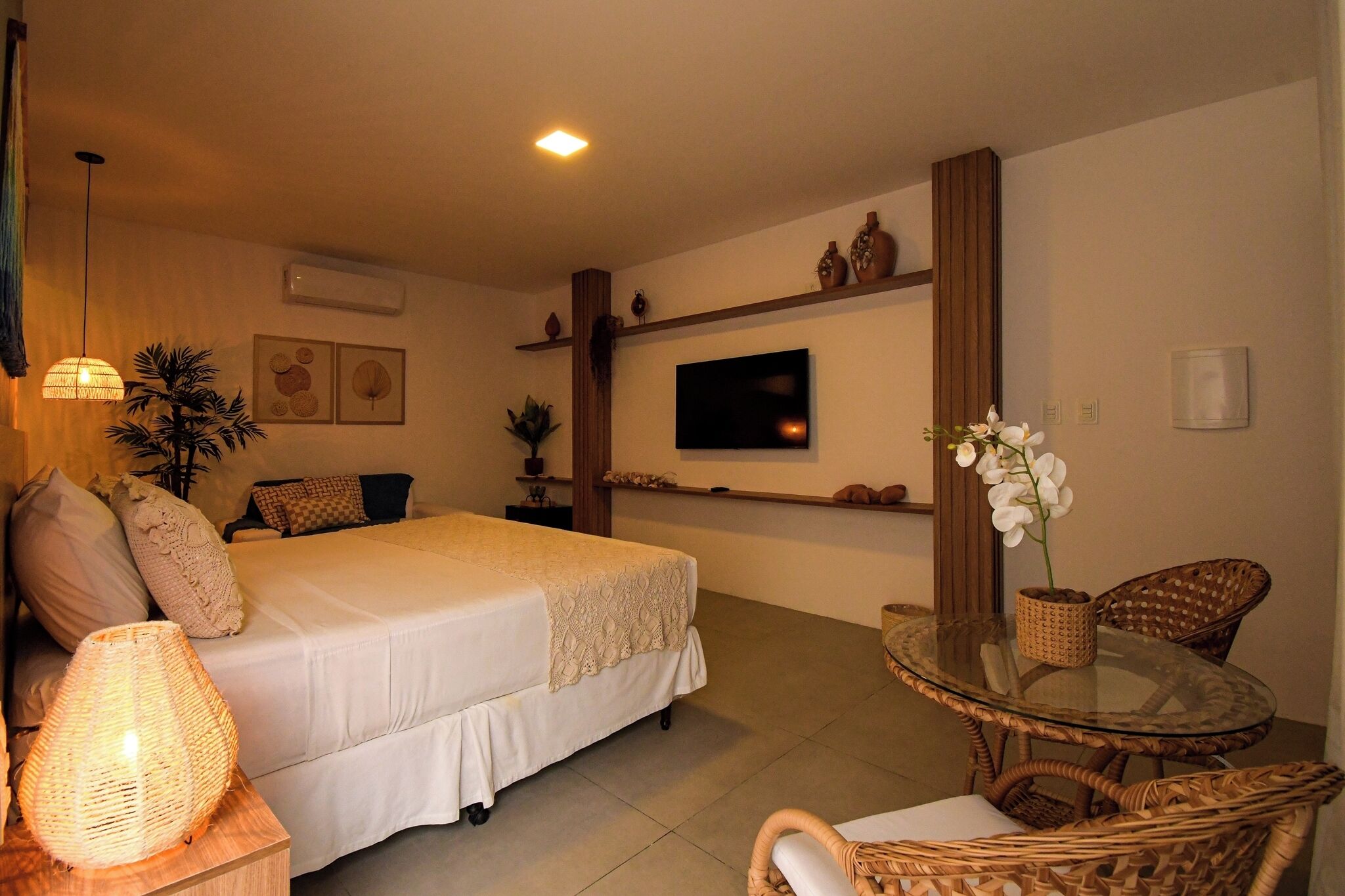 A hotel room with a bed, a glass table with chairs, a TV mounted on the wall, and a potted plant.