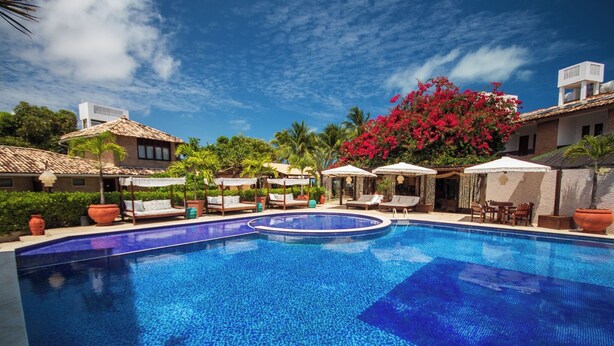 Piscina con sillones de descanso y sombrillas, rodeada de edificios y plantas tropicales.