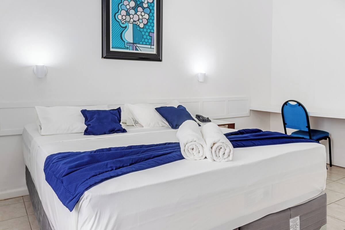 Ein ordentlich bezogenes Bett mit blau-weißer Bettwäsche, ein gerahmtes Kunstwerk an der Wand und ein blauer Sessel.