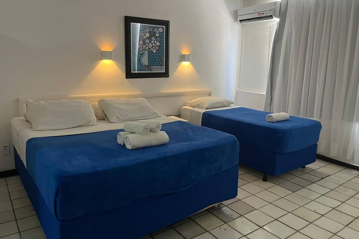 Ein Hotelzimmer mit einem Bett, zwei Nachttischen, einer Wand-Klimaanlage und einem fensterbepflanzten Fenster.