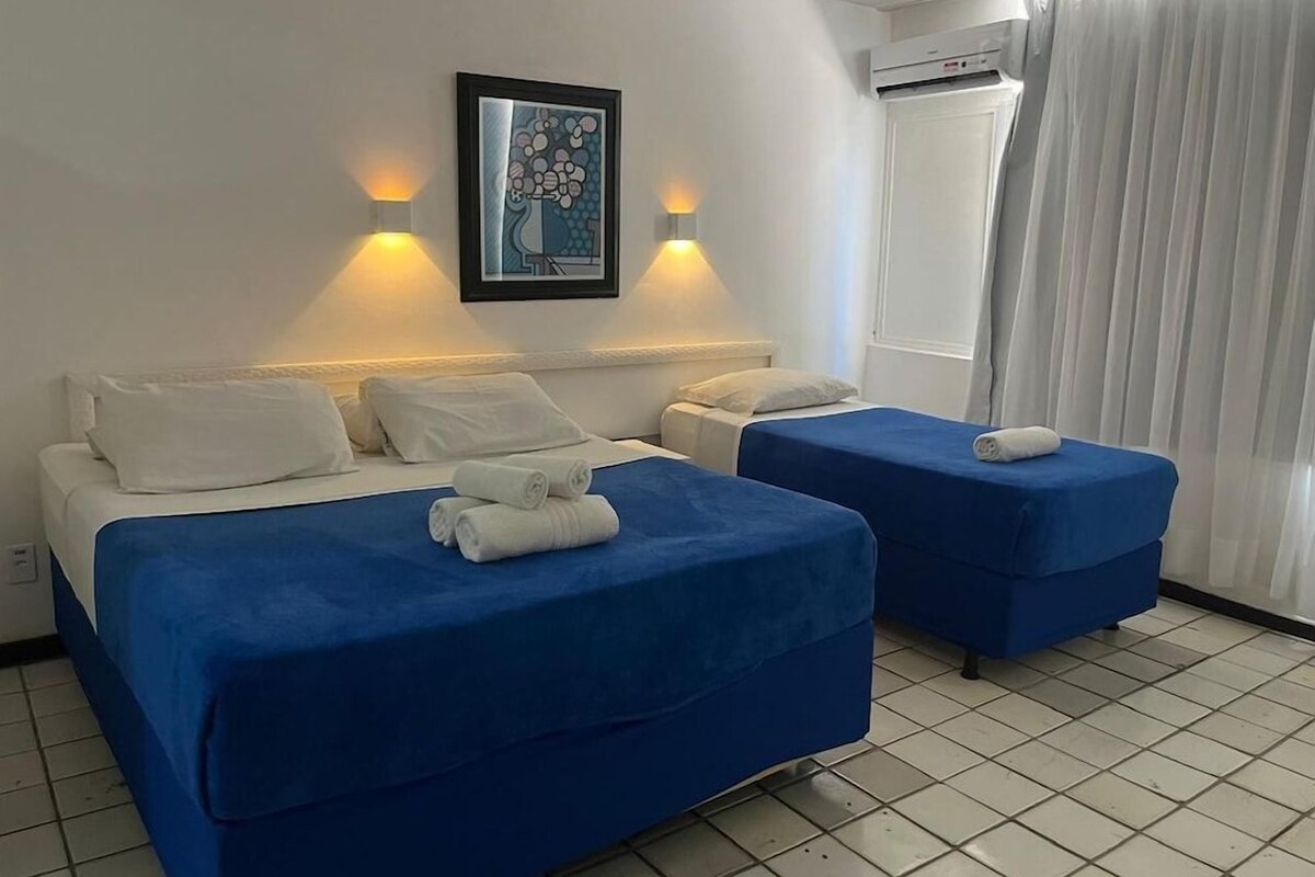 A hotel room with two beds, each with blue bedding and white pillows, and a single bed with white bedding. There is a framed artwork on the wall, a window with curtains, and an air conditioning unit above the window.