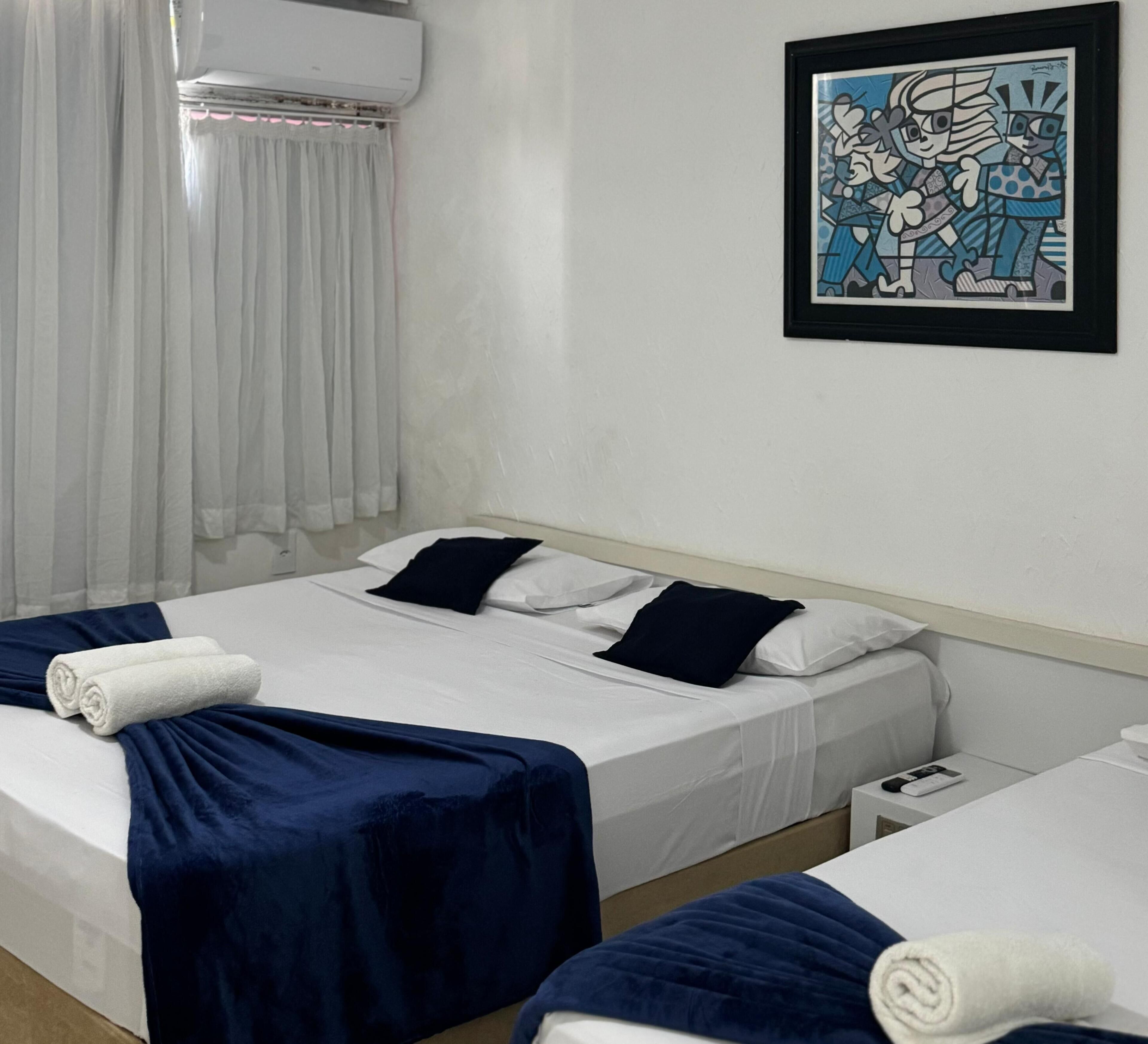 Habitación de hotel con dos camas, sábanas blancas, colchas azul oscuro, almohadas blancas, un cuadro enmarcado en la pared y una cortina blanca.