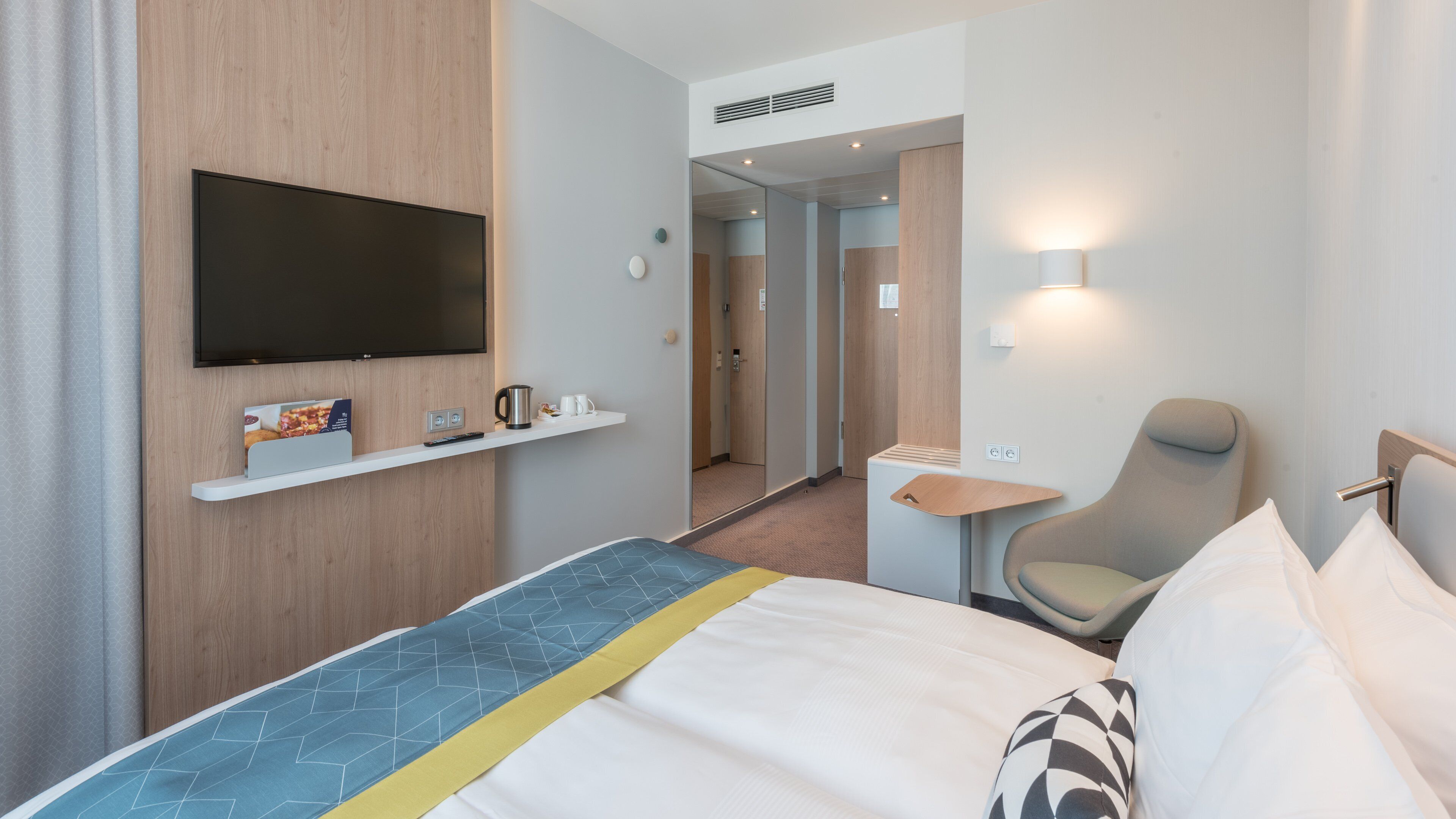 Una habitación de hotel con cama, televisor montado en la pared, una mesita con hervidor y tazas, una silla y un espejo.