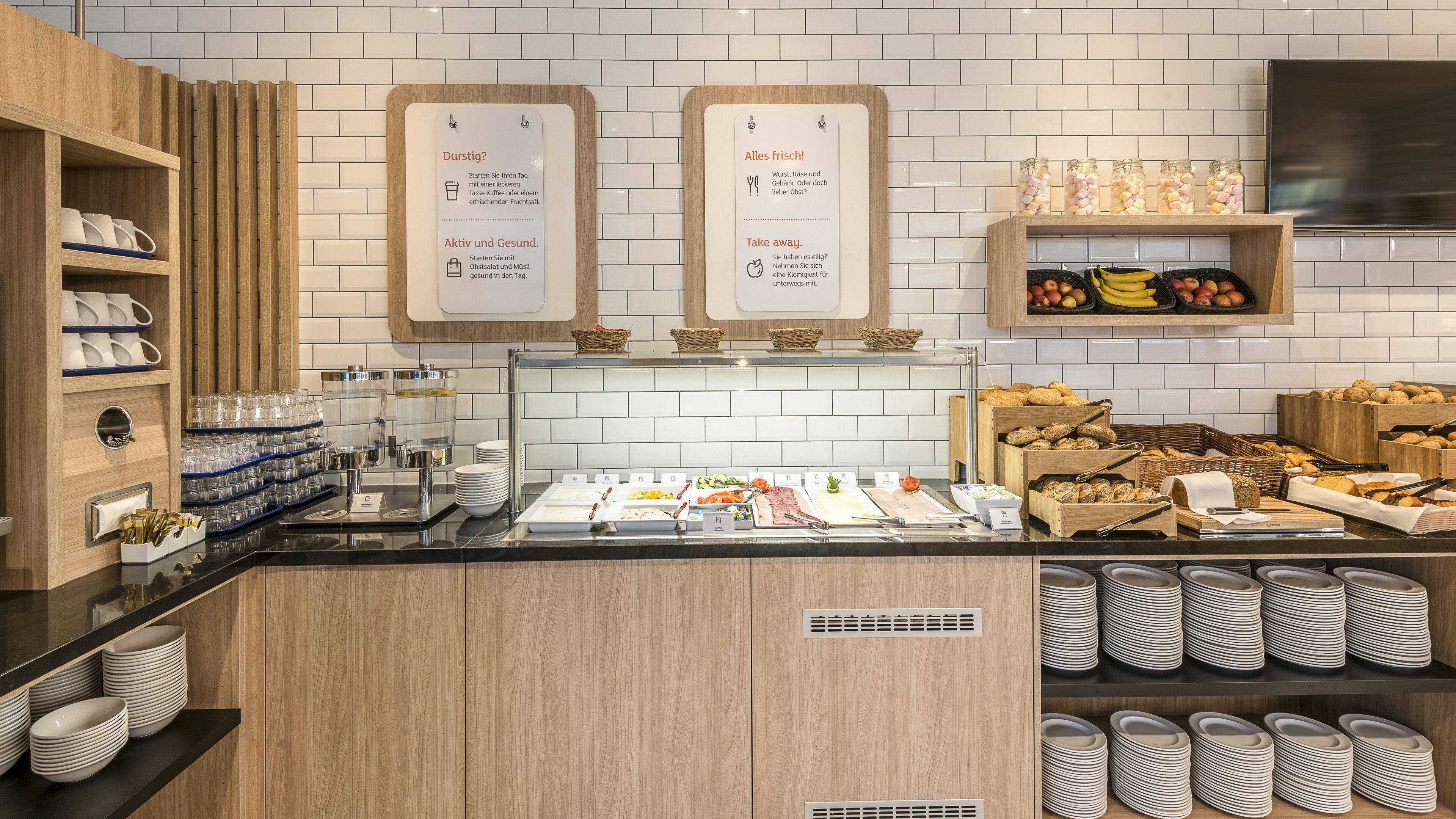 A buffet area with various food options, including fruits, vegetables, and bread. There are signs indicating different sections like 'Durstig?' for drinks and 'Alles frisch!' for fresh food.