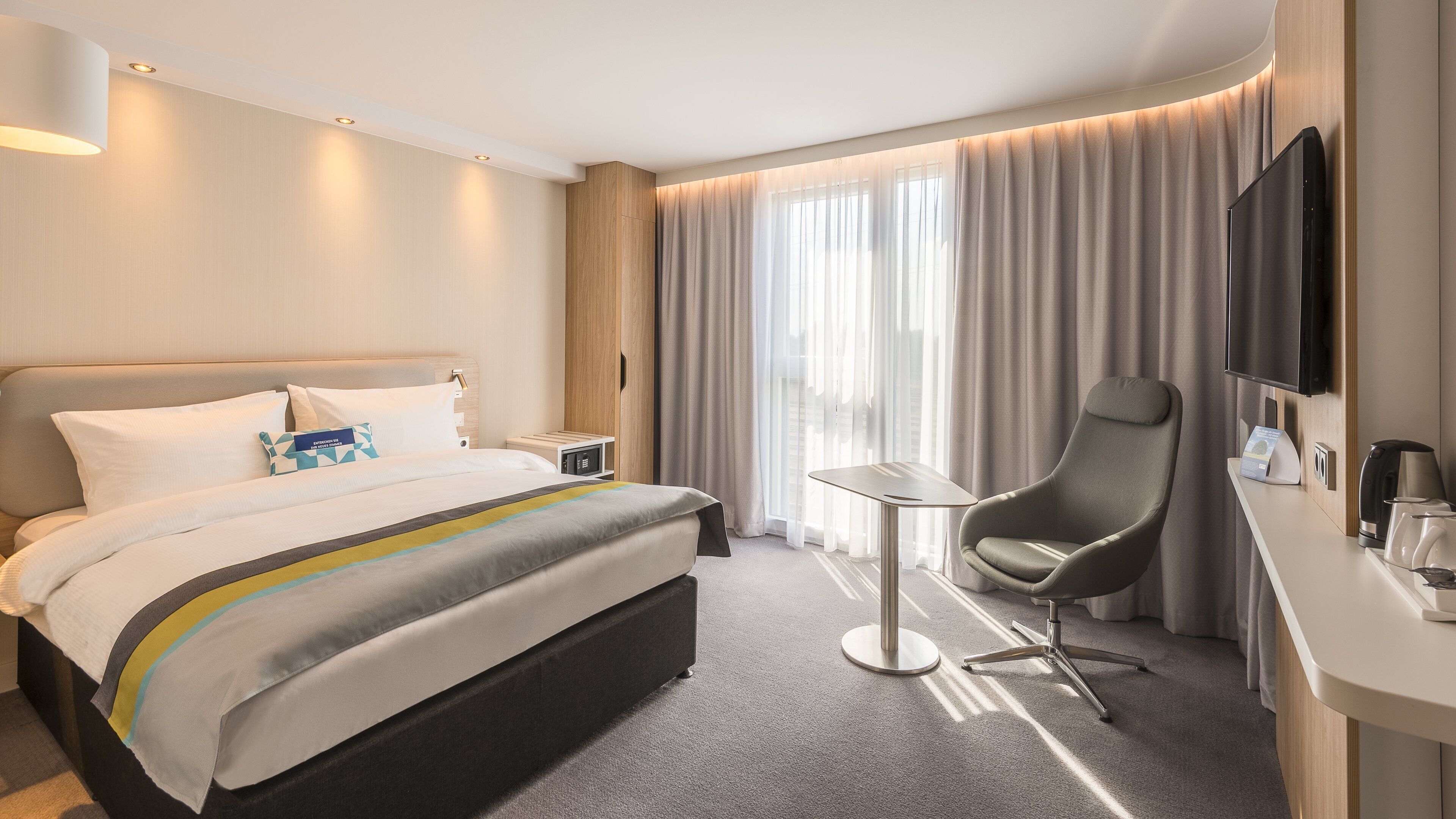 Ein modernes Hotelzimmer mit Bett, Nachttisch, Sessel, Fernseher und Blick auf die Stadt durch große Fenster.