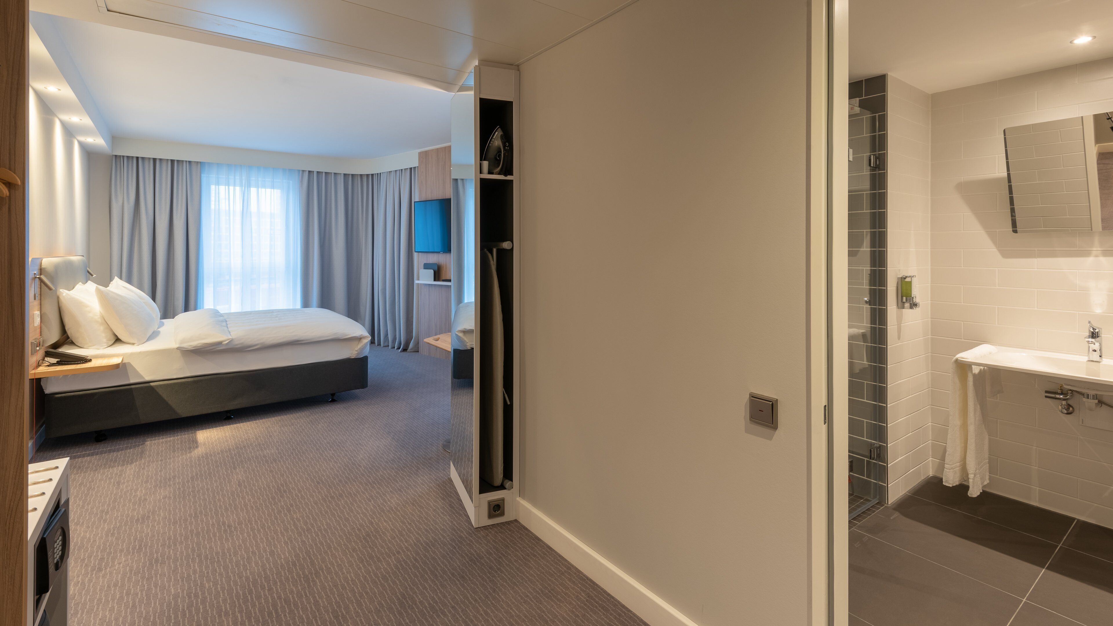Habitación de hotel con cama, escritorio, silla, armario y baño con lavamanos y espejo.