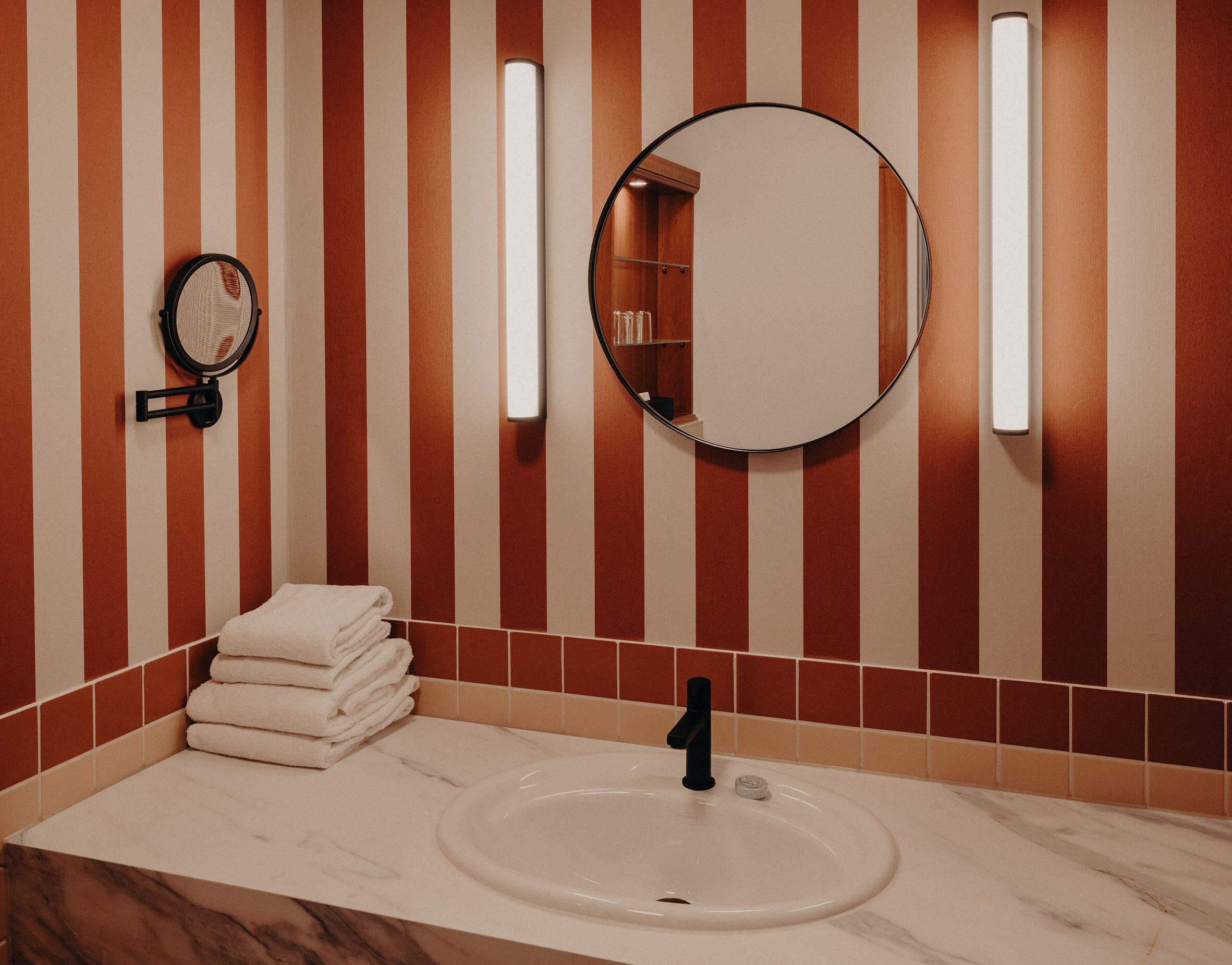 A bathroom with striped walls, a round mirror, a sink with a black faucet, and a stack of white towels.
