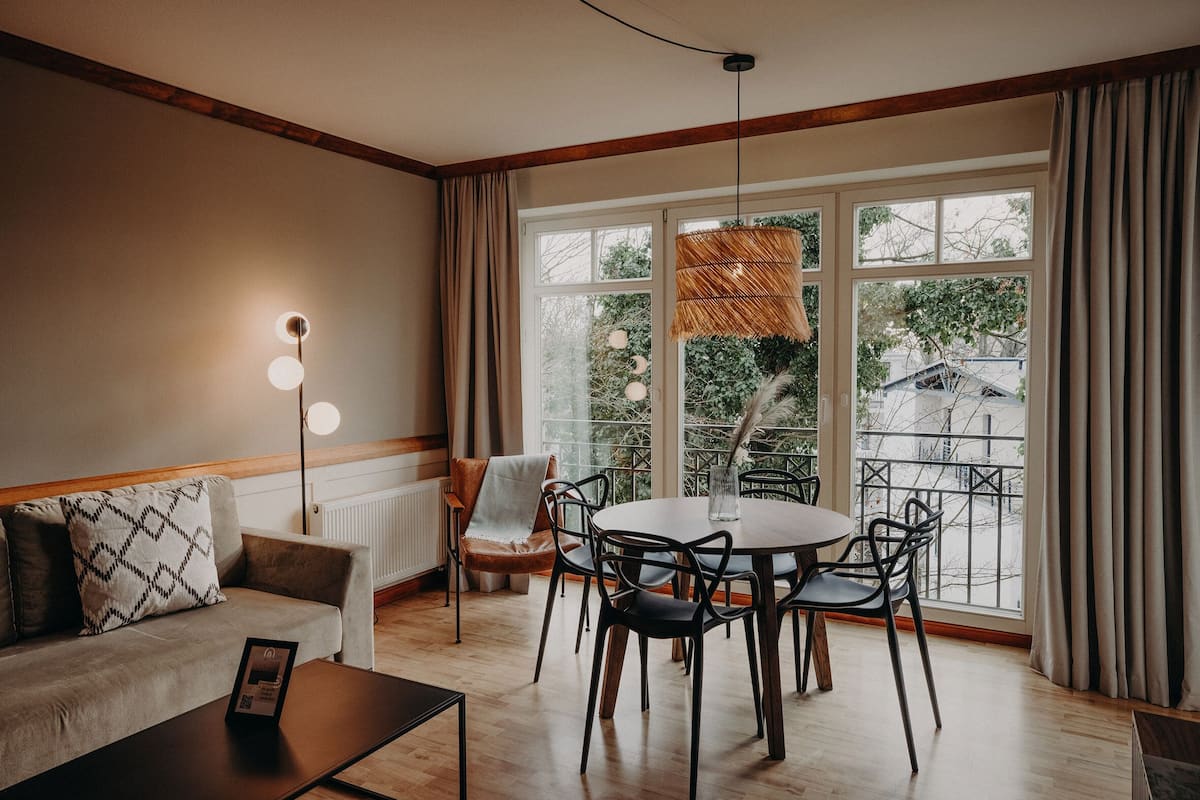 Un salon moderne comprenant un canapé, une table basse et une table à manger avec des chaises. On y trouve une grande fenêtre avec des rideaux, un luminaire suspendu et un radiateur.