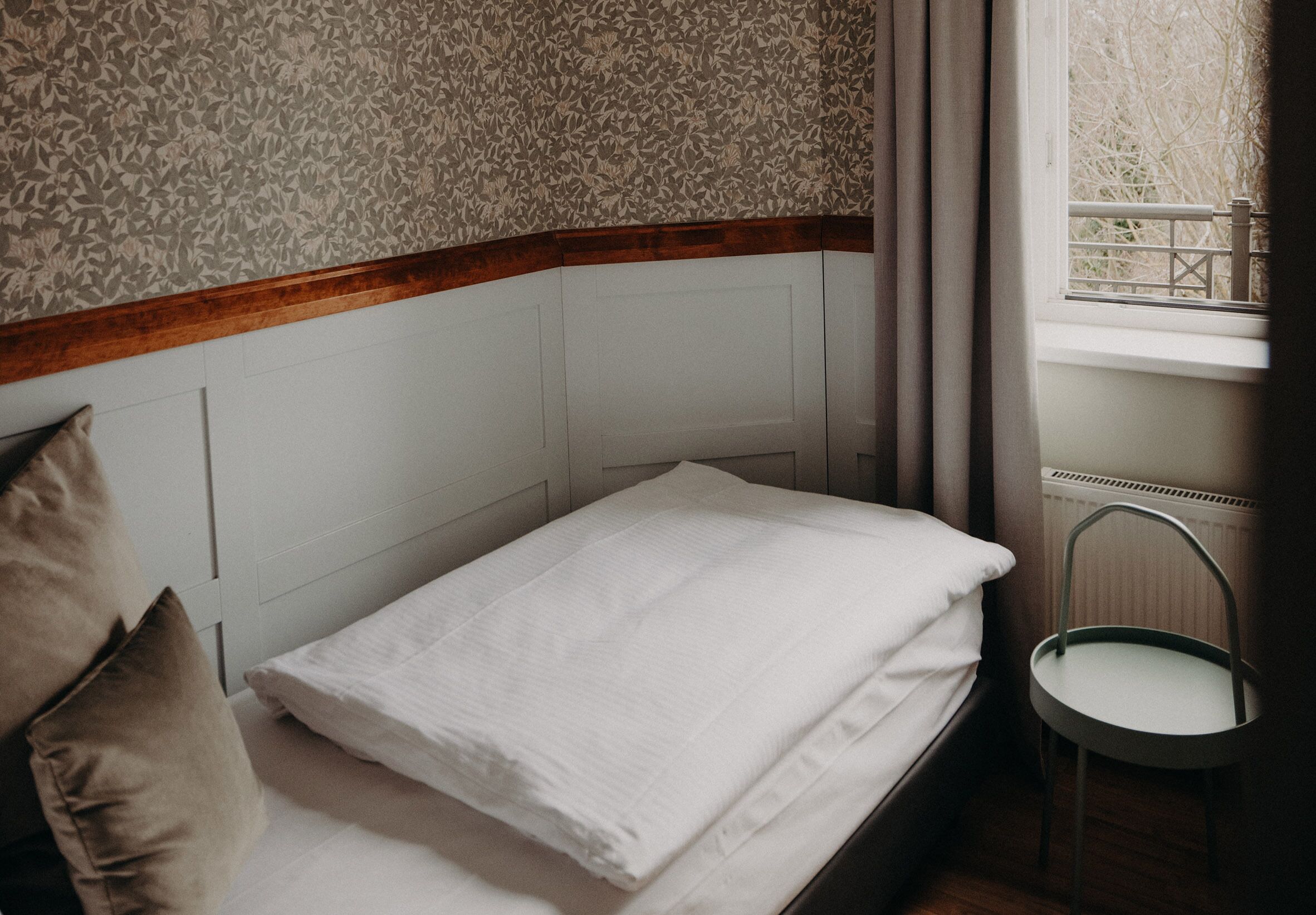 Una cama bien tendida con sábanas blancas en una habitación con papel tapiz floral y cabecera de madera.