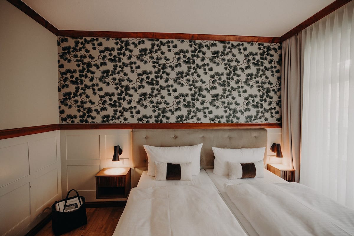Une chambre d’hôtel avec une tête de lit à motifs floraux, un lit double avec du linge de lit blanc, deux lampes de chevet et une table de chevet en bois.