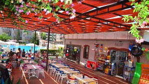 Un café al aire libre con mesas y sillas bajo un toldo rojo, una piscina al fondo y una tienda con diversos artículos a la venta.
