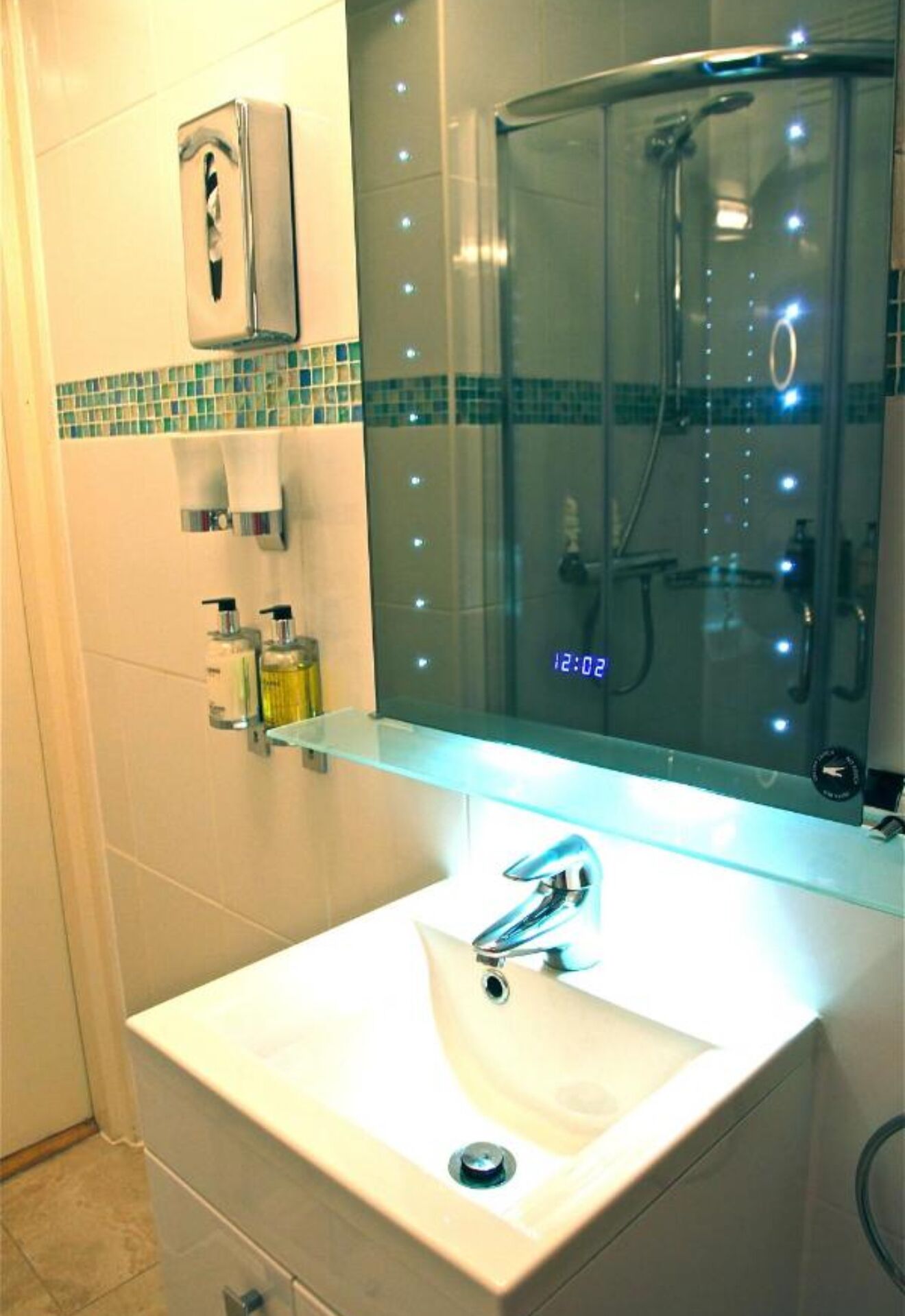 A bathroom with a white sink, a glass shower enclosure, and a mirror with a digital clock displaying 12:02.