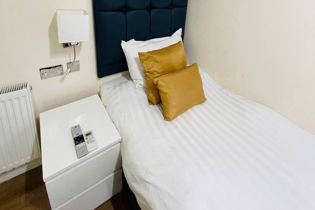A hotel room with a bed, a nightstand, a white bedside table, a wall-mounted air conditioner, and a power outlet.