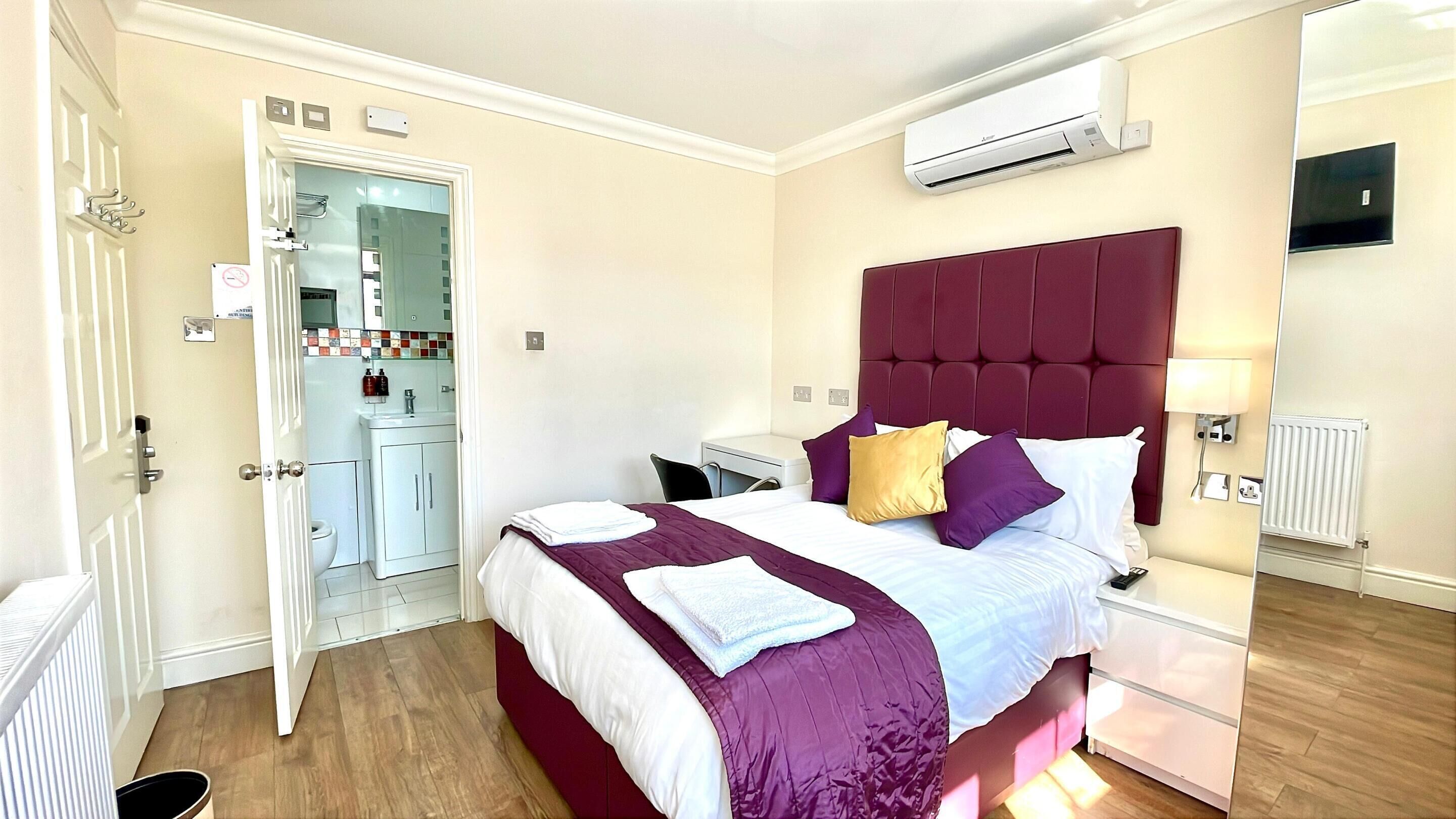 Habitación de hotel con cama, mesitas de noche, aire acondicionado, un espejo y un baño visible a través de una puerta abierta.