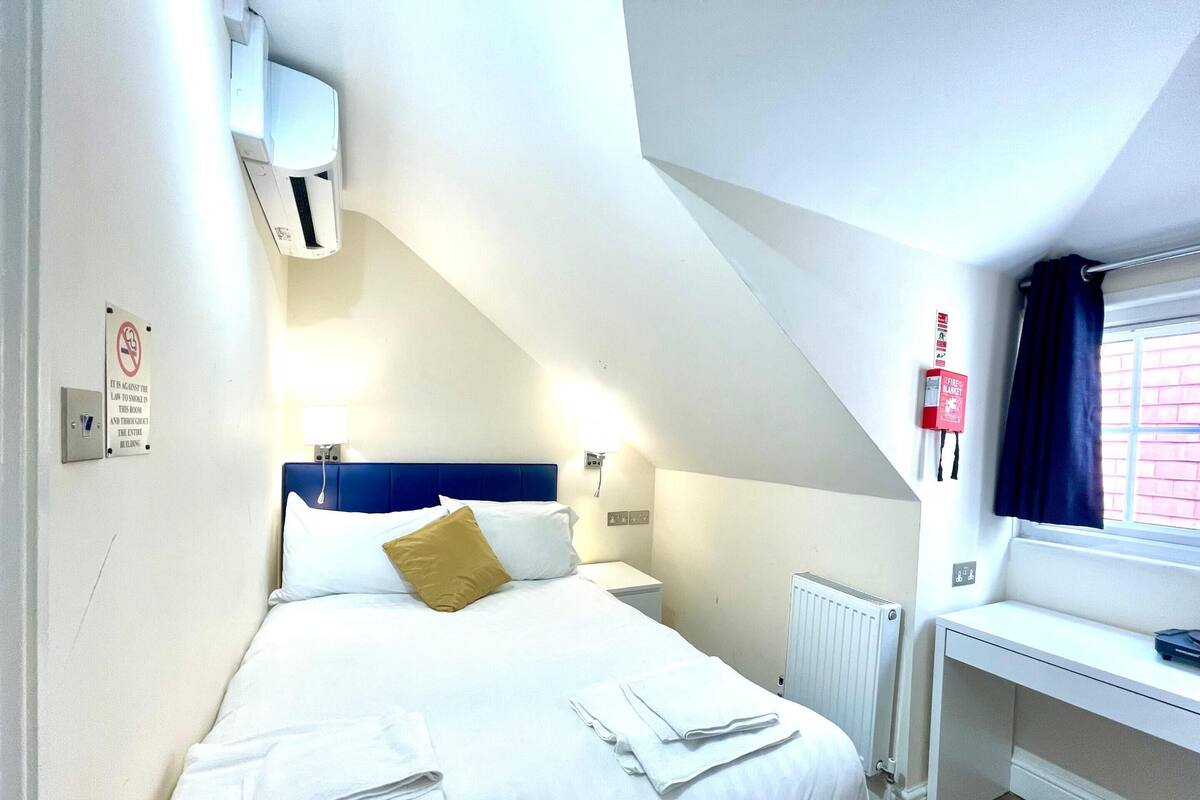 A hotel room with a bed, a desk, a window with curtains, a radiator, and a fire alarm.