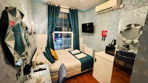 Habitación de hotel pequeña con una cama, un escritorio con silla, un espejo, un televisor y un equipo de aire acondicionado.