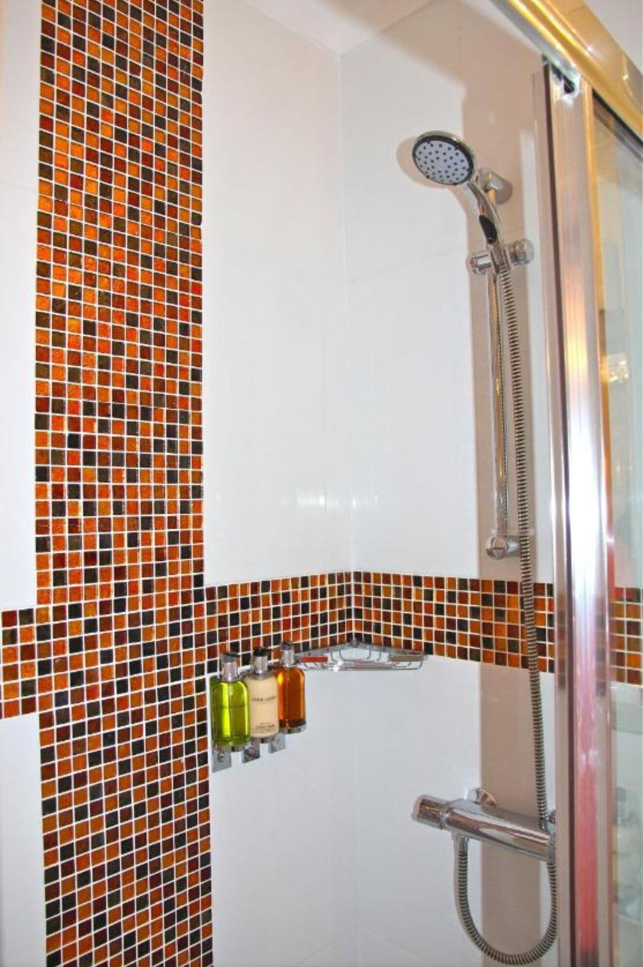 Una ducha con puerta de vidrio, cabezal de ducha de mano y estante con artículos de aseo.