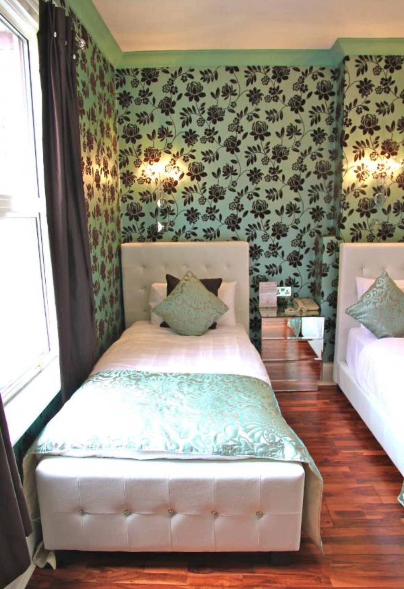 A room with two beds, each with a headboard and a bedside table. The walls have a floral wallpaper pattern, and there is a window with curtains on the left side.