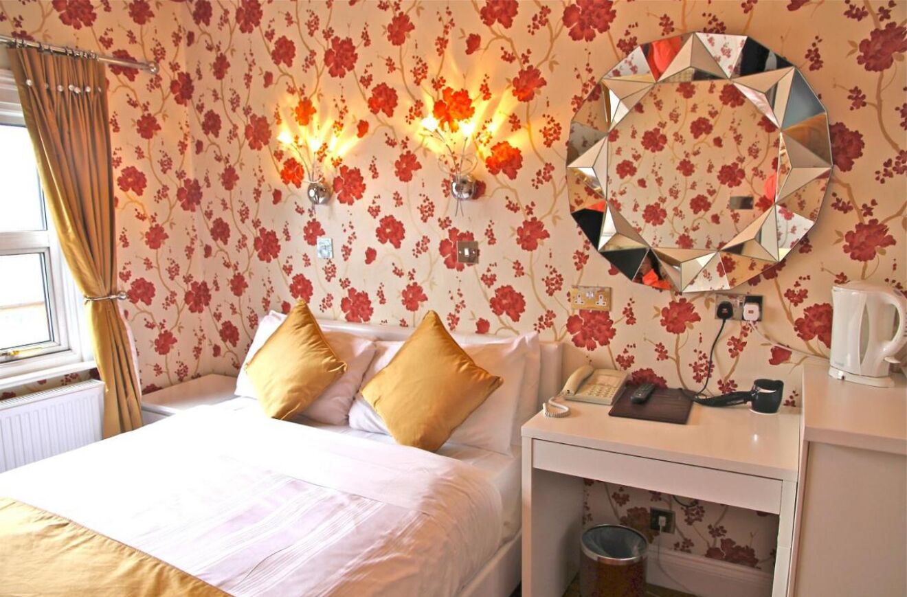 Ein Hotelzimmer mit floralem Wandpapier, einem runden Spiegel, einem Bett mit weißen Leinen, zwei goldenen Kissen, einem Nachttisch mit Telefon, Wasserkocher und Heizkörper.