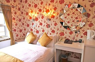 Habitación de hotel con papel tapiz floral, un espejo redondo, una cama con ropa blanca, dos almohadas doradas, una mesita de noche con un teléfono, un hervidor y un radiador.