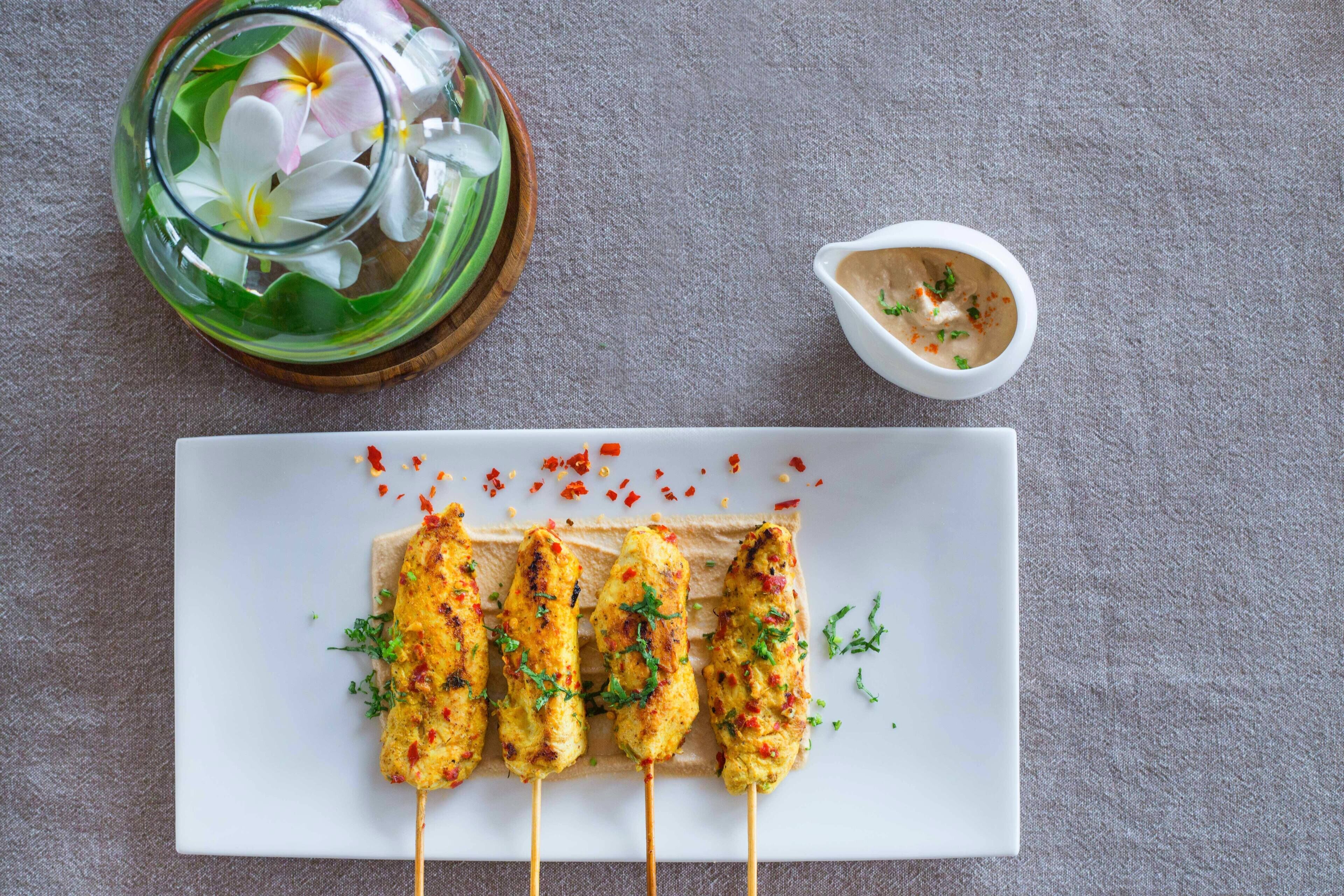 A plate of grilled chicken skewers garnished with herbs, accompanied by a small bowl of sauce and a glass with flowers.