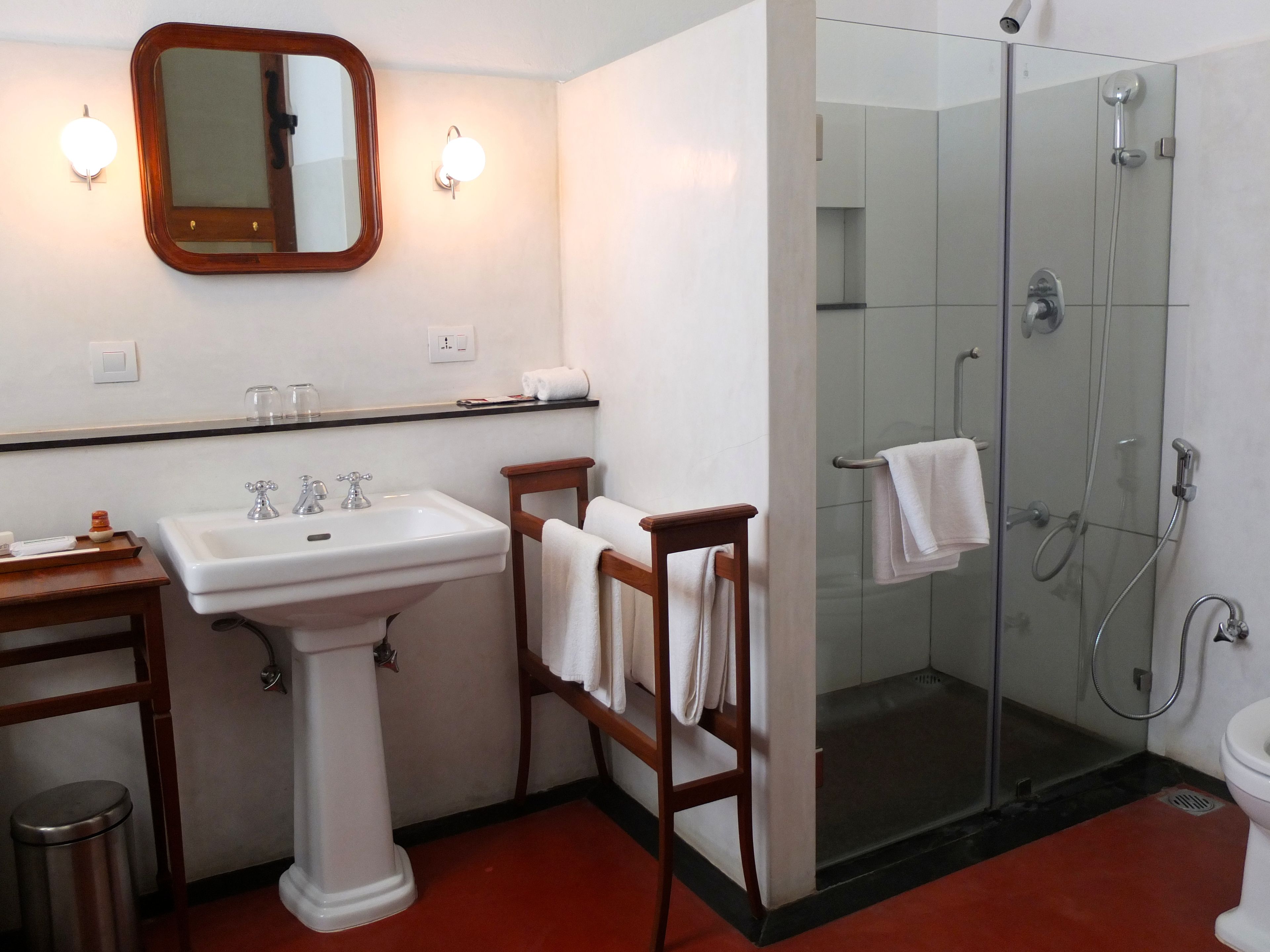 A bathroom with a pedestal sink, a wooden chair with a towel, a glass shower enclosure, and a toilet.